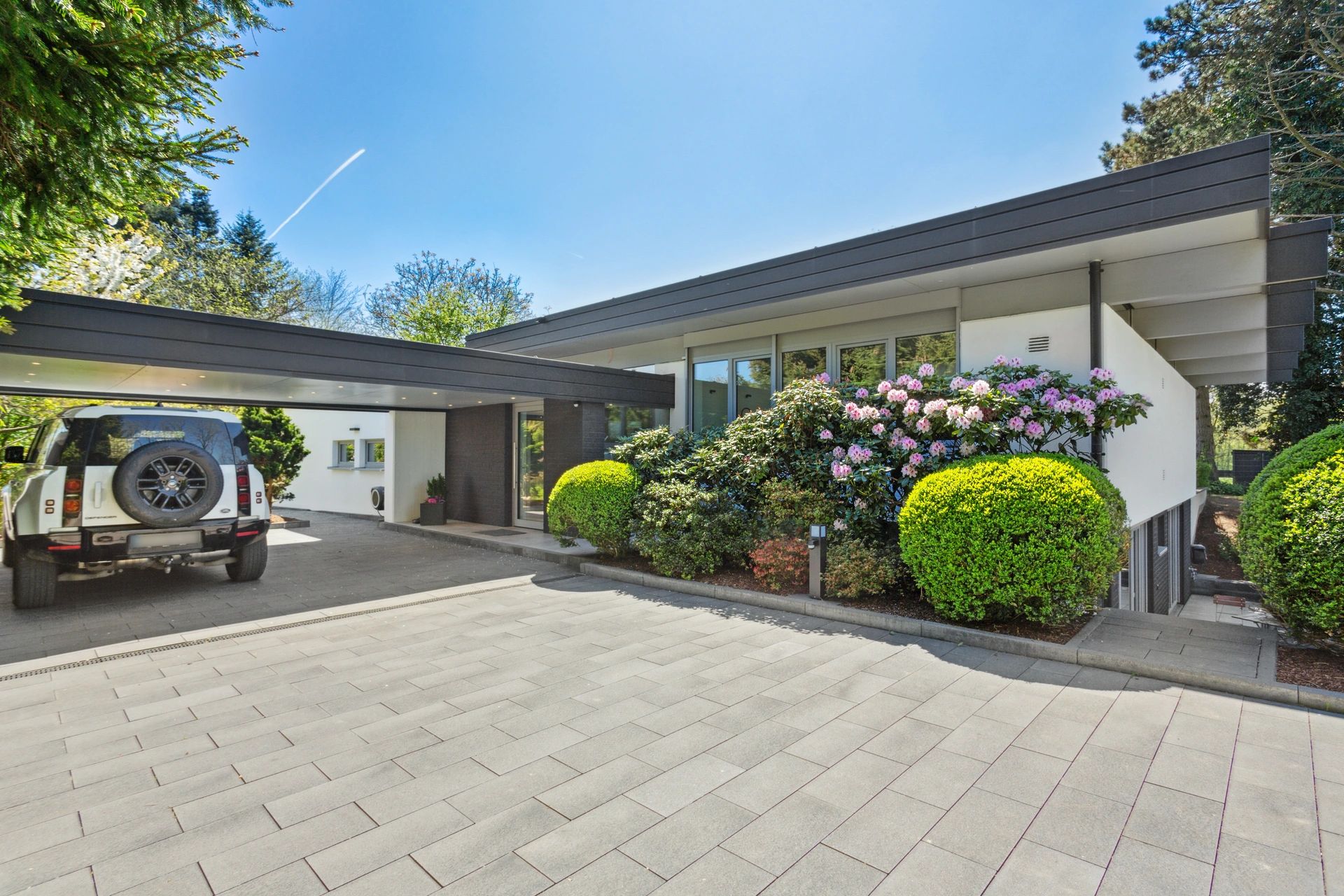 Modernes Flachdachhaus mit Carport, großem Einfahrtsbereich und gepflegtem Vorgarten mit blühenden Rhododendren.
